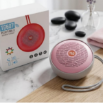 Parlante Bluetooth portátil TG607 circular color rosa con cuerpo gris y correa, junto a su caja blanca con diseño rojo sobre fondo de mármol.