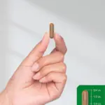 Cápsula marrón translúcida de suplemento de hongos adaptógenos Horbaach, de tres cuartos de pulgada de largo, sostenida con dos dedos junto a una escala de medición verde.
