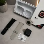 Smartwatch T500 Ultra dorado con dos correas intercambiables, una negra y una gris, cargador magnético, manual y caja sobre superficie gris.