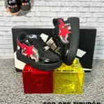 Zapatillas urbanas infantiles negras de charol con diseño de tiburón rojo, suela de goma, exhibidas sobre cubos de colores junto a su caja.