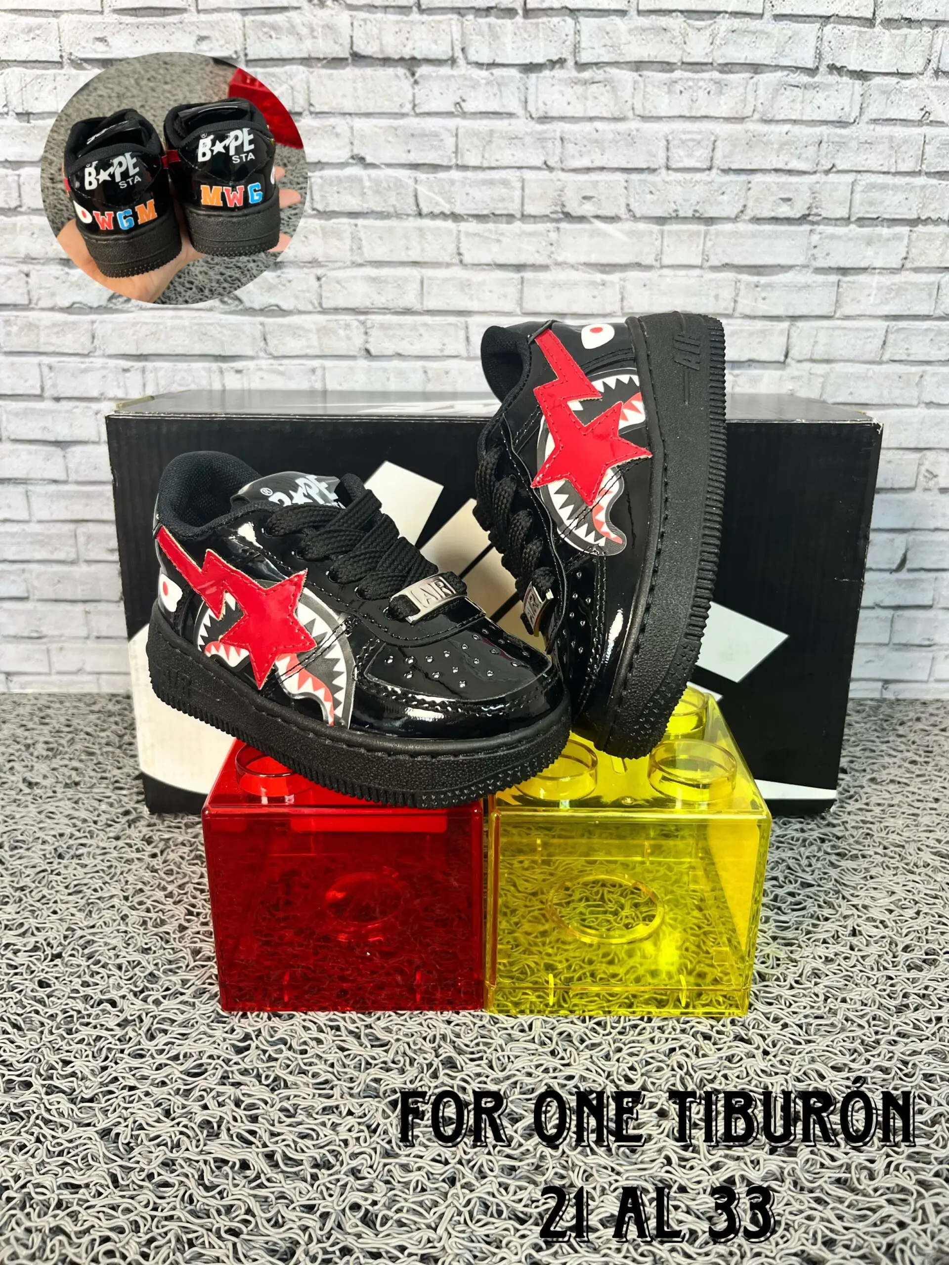 Zapatillas urbanas infantiles negras de charol con diseño de tiburón rojo, suela de goma, exhibidas sobre cubos de colores junto a su caja.