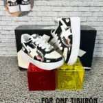 Zapatillas urbanas infantiles negras y blancas con diseño de tiburón en charol, expuestas sobre bloques de plástico rojo y amarillo ante una caja y fondo de ladrillo, tallas 21 a 33.