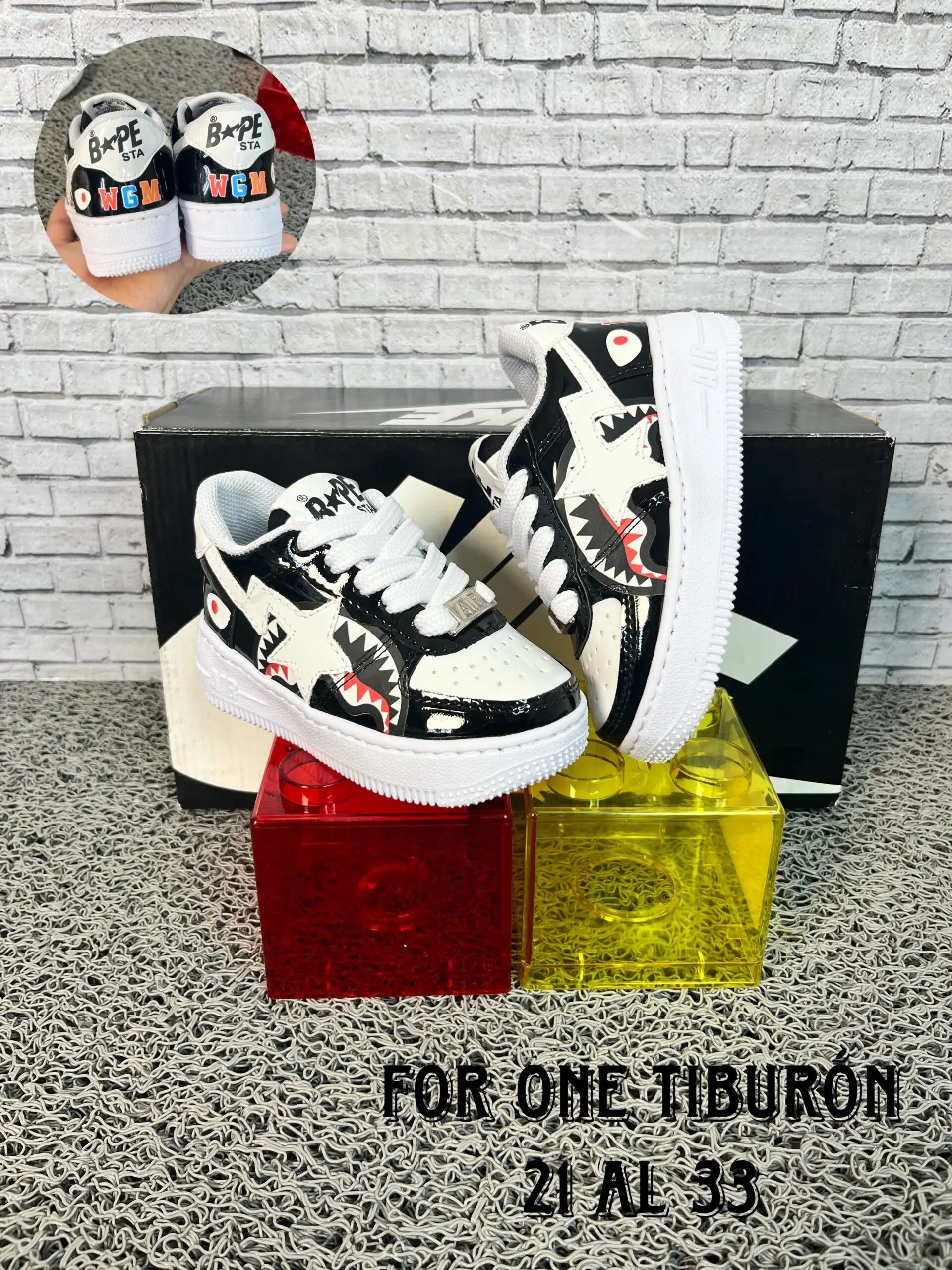 Zapatillas urbanas infantiles negras y blancas con diseño de tiburón en charol, expuestas sobre bloques de plástico rojo y amarillo ante una caja y fondo de ladrillo, tallas 21 a 33.