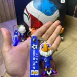 Llavero azul de la Copa Mundial con mascota de águila en uniforme deportivo y balón, sostenido en primer plano con empaque al fondo.