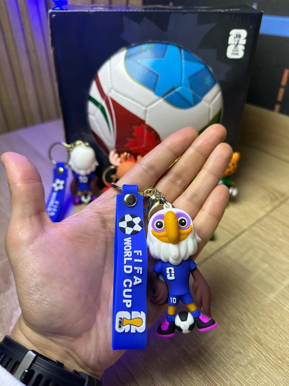 Llavero azul de la Copa Mundial con mascota de águila en uniforme deportivo y balón, sostenido en primer plano con empaque al fondo.