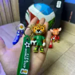 Llavero de mascota mundialista leopardo en verde con correa de FIFA World Cup, junto a variantes azul y roja y caja del set de fútbol.