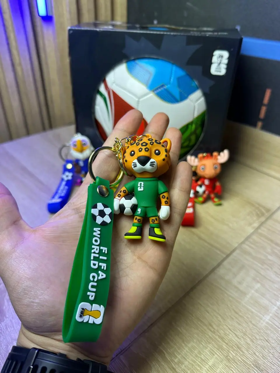 Llavero de mascota mundialista leopardo en verde con correa de FIFA World Cup, junto a variantes azul y roja y caja del set de fútbol.