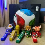 Set de llaveros de fútbol con figuras de mascotas y copa dorada en colores azul, amarillo, verde y rojo, frente a un balón en caja negra.