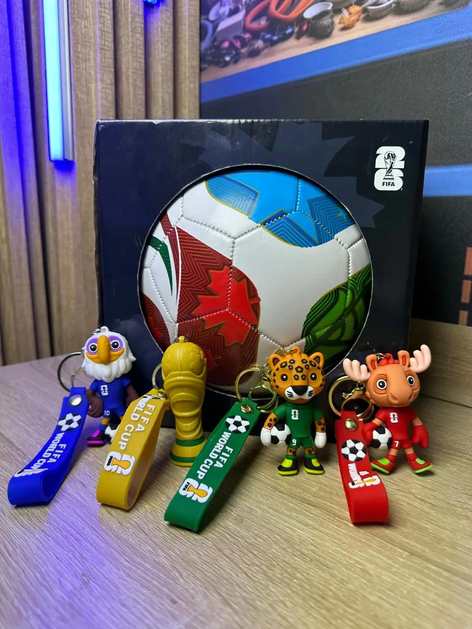 Set de llaveros de fútbol con figuras de mascotas y copa dorada en colores azul, amarillo, verde y rojo, frente a un balón en caja negra.