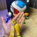 Llavero de silicona amarillo con trofeo dorado y correa FIFA World Cup, junto a figuras de mascotas y caja temática sobre superficie de madera.