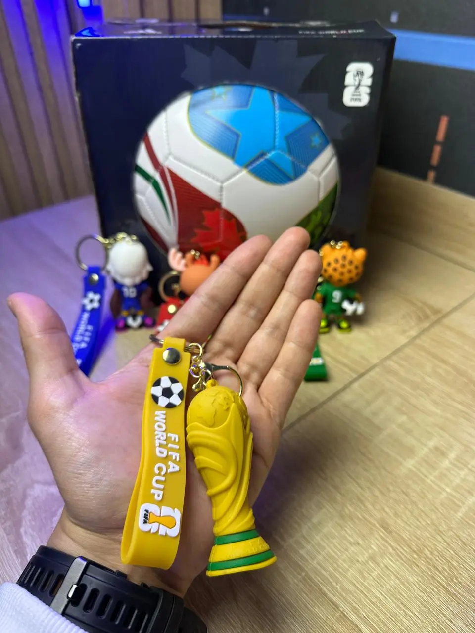 Llavero de silicona amarillo con trofeo dorado y correa FIFA World Cup, junto a figuras de mascotas y caja temática sobre superficie de madera.