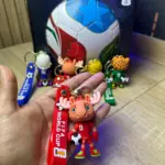 Llaveros de fútbol con mascotas mundialistas. Mano sostiene modelo rojo de alce con correa FIFA World Cup; al fondo balón y variantes de colores.