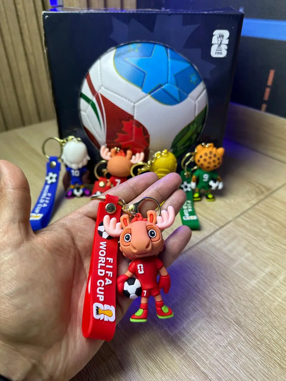 Llaveros de fútbol con mascotas mundialistas. Mano sostiene modelo rojo de alce con correa FIFA World Cup; al fondo balón y variantes de colores.