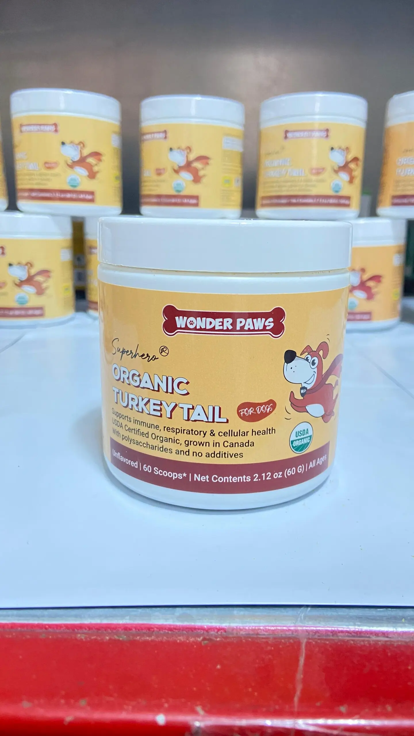 Tarro blanco con etiqueta naranja de suplemento en polvo de hongo cola de pavo para perros, presentación de 60 gramos, ubicado en estantería con otros envases idénticos.