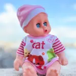 Muñeca bebé de juguete con ojos azules, gorro rosa y mameluco blanco con rayas y dibujos de animales, sentada sobre piedra bajo cielo azul.