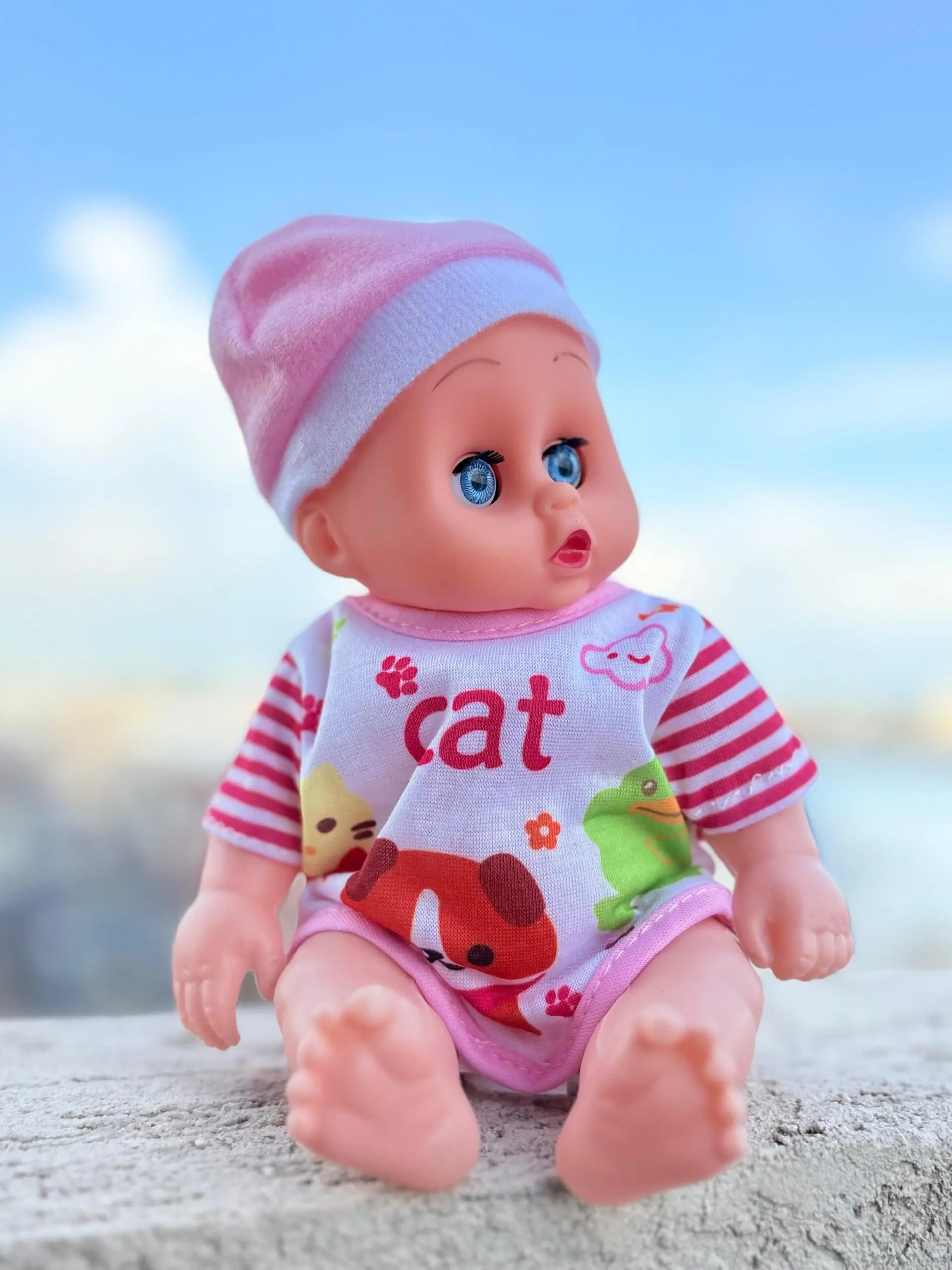 Muñeca bebé de juguete con ojos azules, gorro rosa y mameluco blanco con rayas y dibujos de animales, sentada sobre piedra bajo cielo azul.