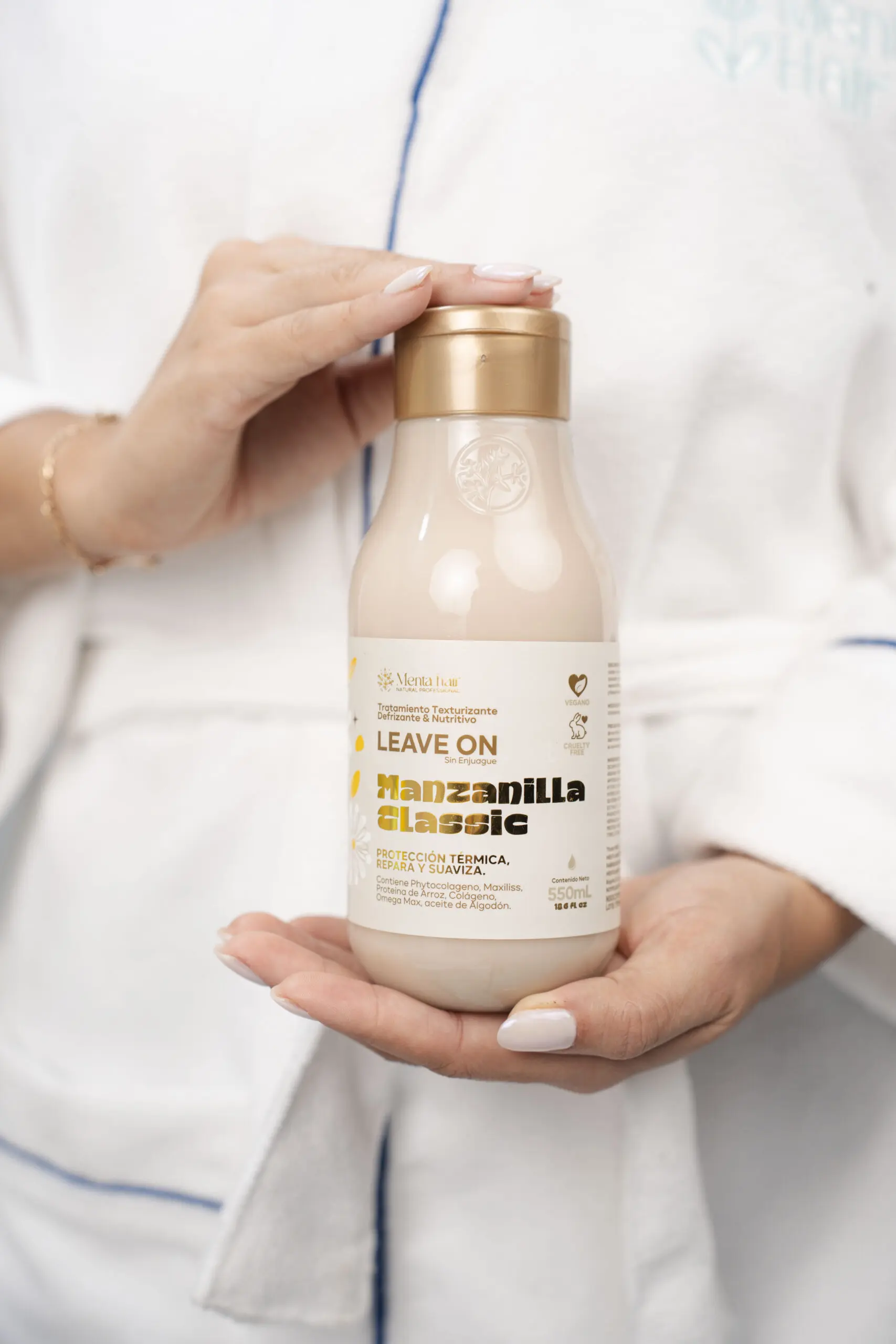 Manos sosteniendo un frasco de tratamiento capilar Manzanilla Classic de 550ml, con fórmula termoprotectora y anti frizz, en envase beige y dorado.