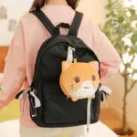 Mochila escolar negra de nailon con correas acolchadas, lazos laterales rosados y un peluche colgante en forma de gato naranja.