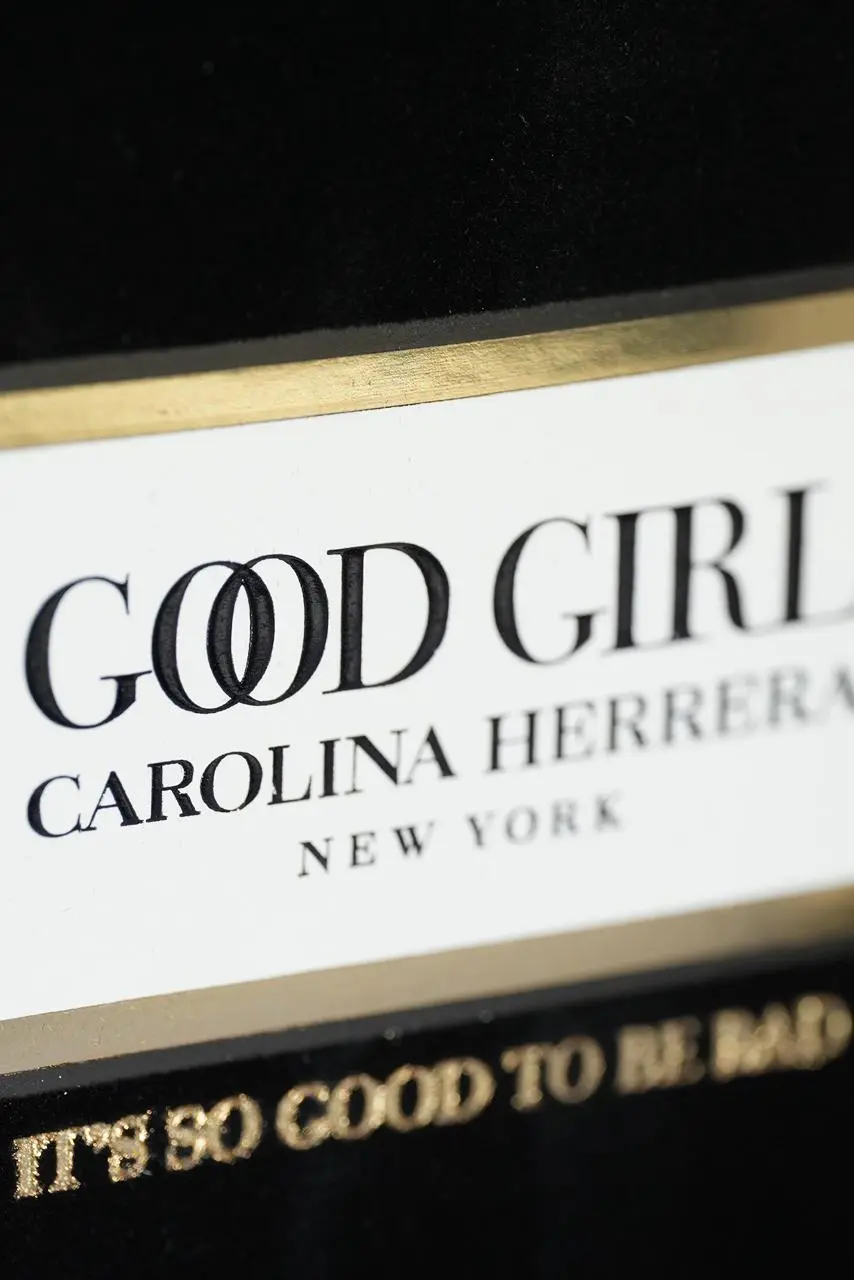 Detalle cercano de la etiqueta en relieve dorada y blanca del perfume Good Girl de Carolina Herrera, sobre superficie de terciopelo negro.