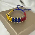 Manilla doble de neopreno con cuentas amarillas, azules y rojas intercaladas con esferas de oro de 18k, sobre una caja café.