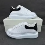 Tenis Alexander McQueen blancos de cuero con suela plataforma y detalle de brillo negro en el talón, sobre una caja blanca.
