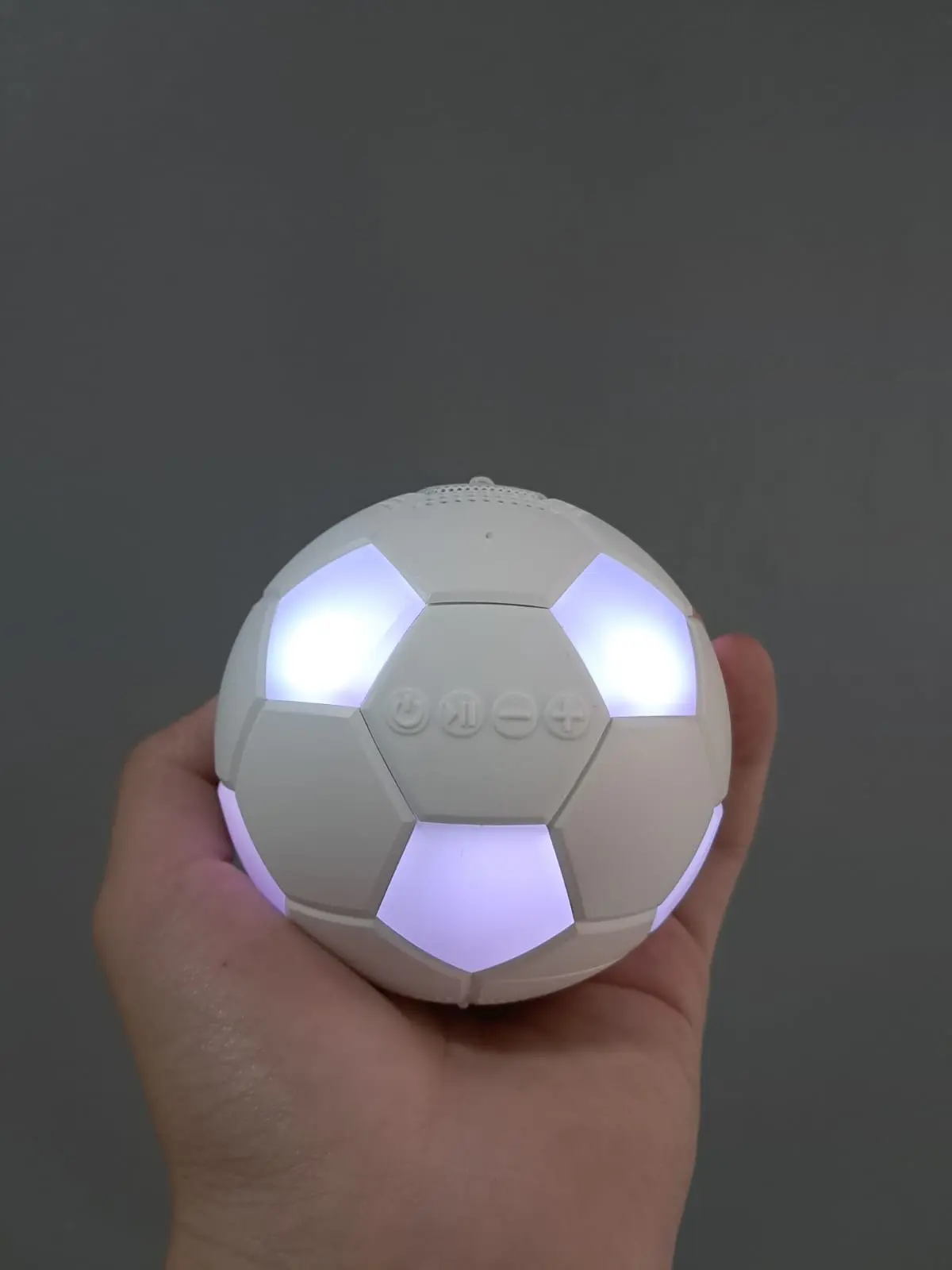 Mano sosteniendo parlante inalámbrico en forma de balón blanco con luces LED brillantes integradas en sus paneles y botones de control visibles.