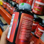 Frasco negro de suplemento de arándano rojo Greensoft con etiqueta roja, sostenido en mano frente a estantes de productos en una tienda.