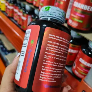 Frasco negro de suplemento de arándano rojo Greensoft con etiqueta roja, sostenido en mano frente a estantes de productos en una tienda.