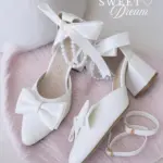 Zapatos de tacón en bloque blancos con perlas y lazo satinado, sobre alfombra rosada.