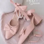Zapatos de tacón en bloque color rosa claro, acabado brillante, con lazos decorativos en la punta y perlas colgantes en el tobillo.