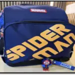 Morral escolar azul marino con letras amarillas de Spiderman, logo de Marvel y un llavero de Spiderman colgando del cierre frontal.