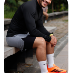 Hombre sonriente usando buso deportivo negro de manga larga, pantaloneta gris y tenis naranjas, sentado al aire libre.