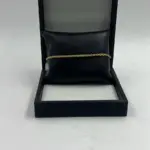 Pulsera dorada delgada con diseño de cuerda trenzada, expuesta horizontalmente sobre un cojín de cuero sintético negro dentro de un estuche abierto.