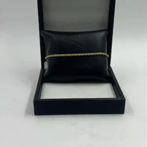 Pulsera dorada delgada con diseño de cuerda trenzada, expuesta horizontalmente sobre un cojín de cuero sintético negro dentro de un estuche abierto.