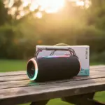 Parlante portátil inalámbrico negro con luces LED multicolor en los extremos, asa superior y caja de empaque sobre mesa de madera al aire libre.