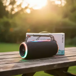 Parlante portátil inalámbrico negro con luces LED multicolor en los extremos, asa superior y caja de empaque sobre mesa de madera al aire libre.