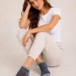 Mujer sentada usando medias tobilleras grises de tejido suave con corazones coloridos y rayas en la punta, con top blanco y pantalón claro.