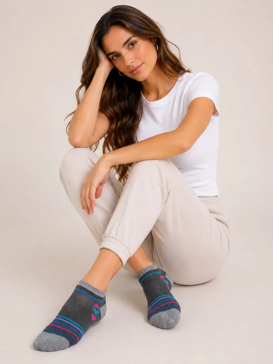 Mujer sentada usando medias tobilleras grises de tejido suave con corazones coloridos y rayas en la punta, con top blanco y pantalón claro.
