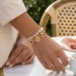 Pulsera de cuarzo rosa con dijes dorados colgantes, corazón y cruz en brazo de mujer tomando café.