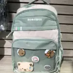 Morral verde y beige para portátil con diseño casual, bolsillo frontal de malla con pines decorativos y oso de peluche aplicado.