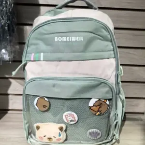 Morral verde y beige para portátil con diseño casual, bolsillo frontal de malla con pines decorativos y oso de peluche aplicado.