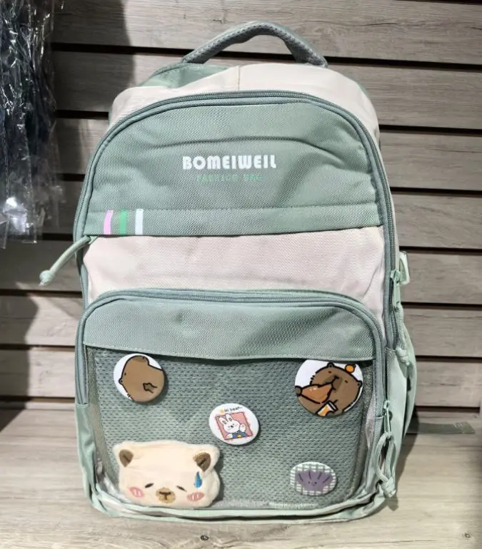 Morral verde y beige para portátil con diseño casual, bolsillo frontal de malla con pines decorativos y oso de peluche aplicado.