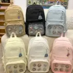 Seis morrales urbanos de lona en tonos beige, negro, azul claro, amarillo, blanco y rosa, con bolsillo frontal transparente lleno de pines decorativos.