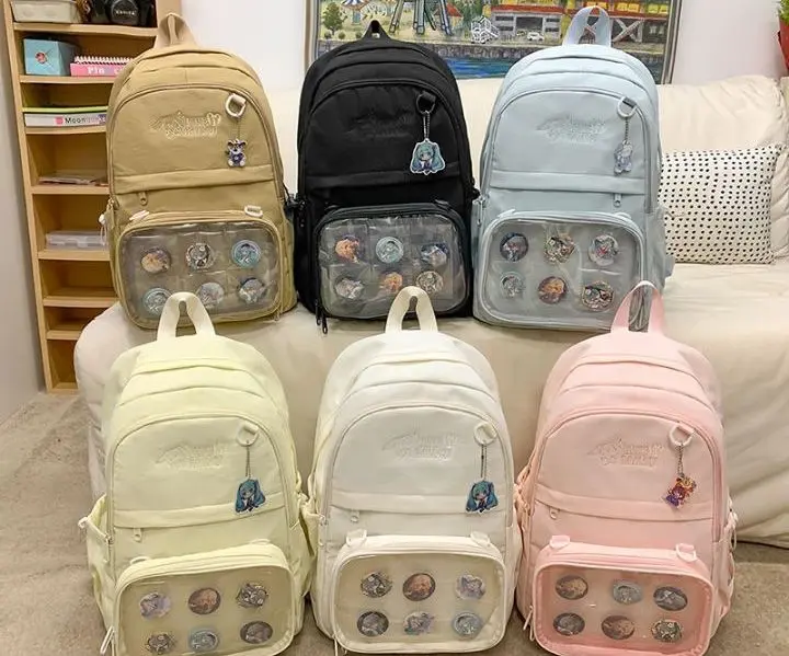 Seis morrales urbanos de lona en tonos beige, negro, azul claro, amarillo pastel, blanco y rosado, con bolsillo frontal transparente lleno de pines decorativos.