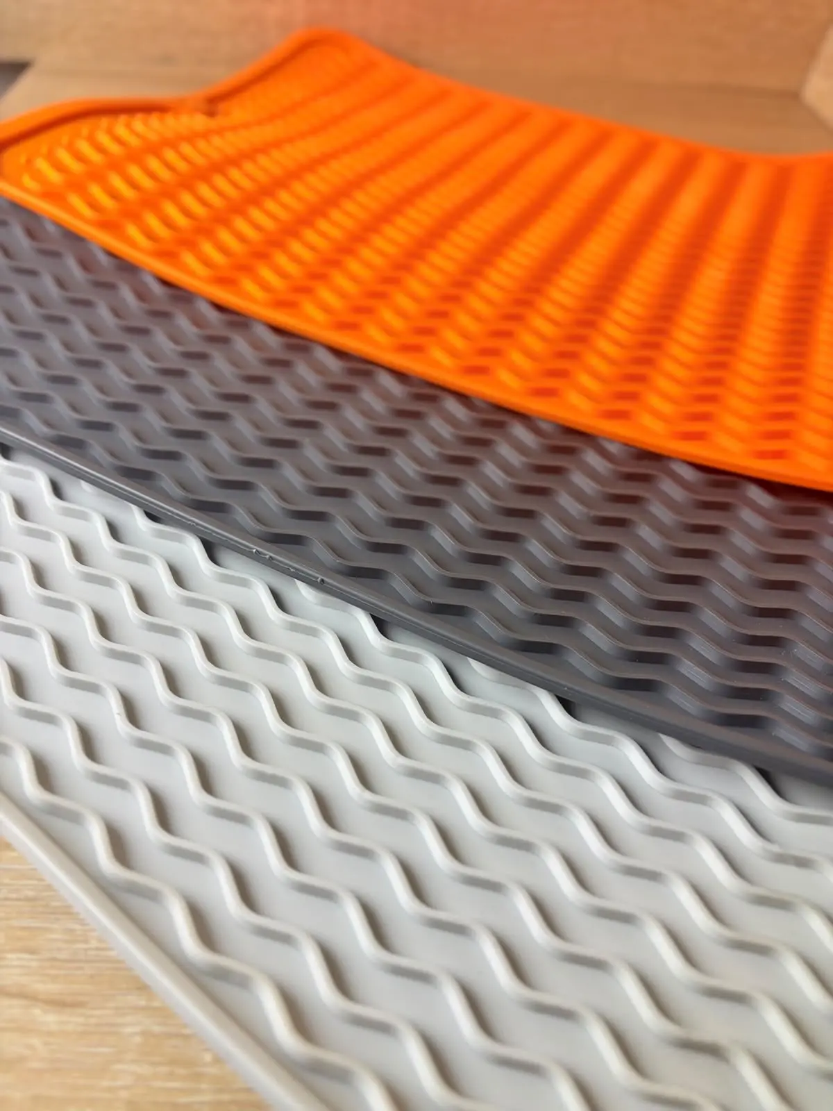 Apilamiento de tres tapetes de silicona antideslizantes para cocina en colores naranja, gris oscuro y gris claro con relieve ondulado térmico.