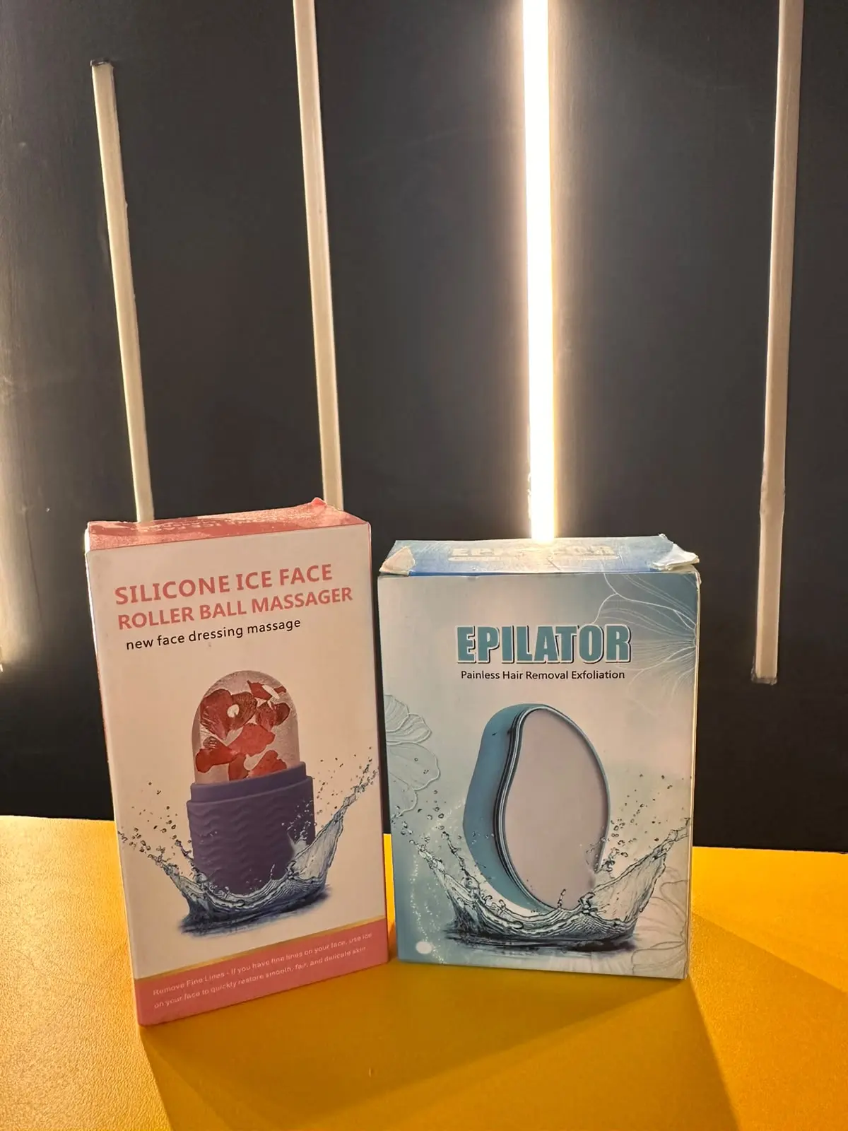 Empaques de rodillo facial de silicona con pétalos de rosa y depilador de nanocristal en tonos rosa y azul sobre superficie amarilla.