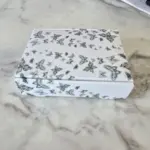 Caja blanca de regalo con estampado de mariposas en tonos grises sobre superficie de mármol.