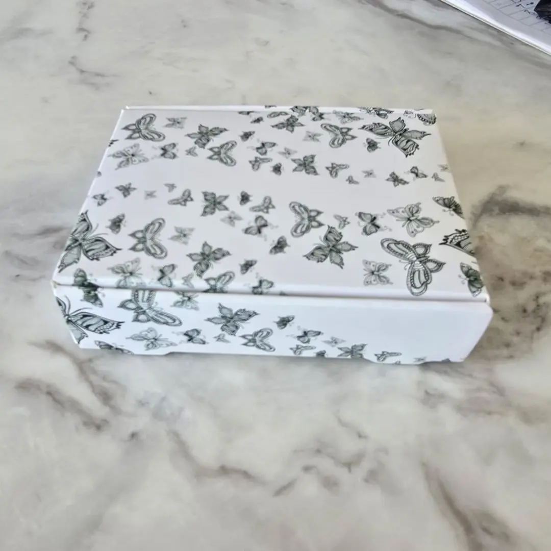 Caja blanca de regalo con estampado de mariposas en tonos grises sobre superficie de mármol.
