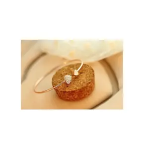 Pulsera dorada rosada con abertura y dos dijes de corazón, uno liso y otro pavé, apoyada sobre un cilindro de corcho.