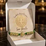 Pulsera tejida verde con cuentas y medalla dorada de San Benito dentro de caja blanca con tarjeta de protección.